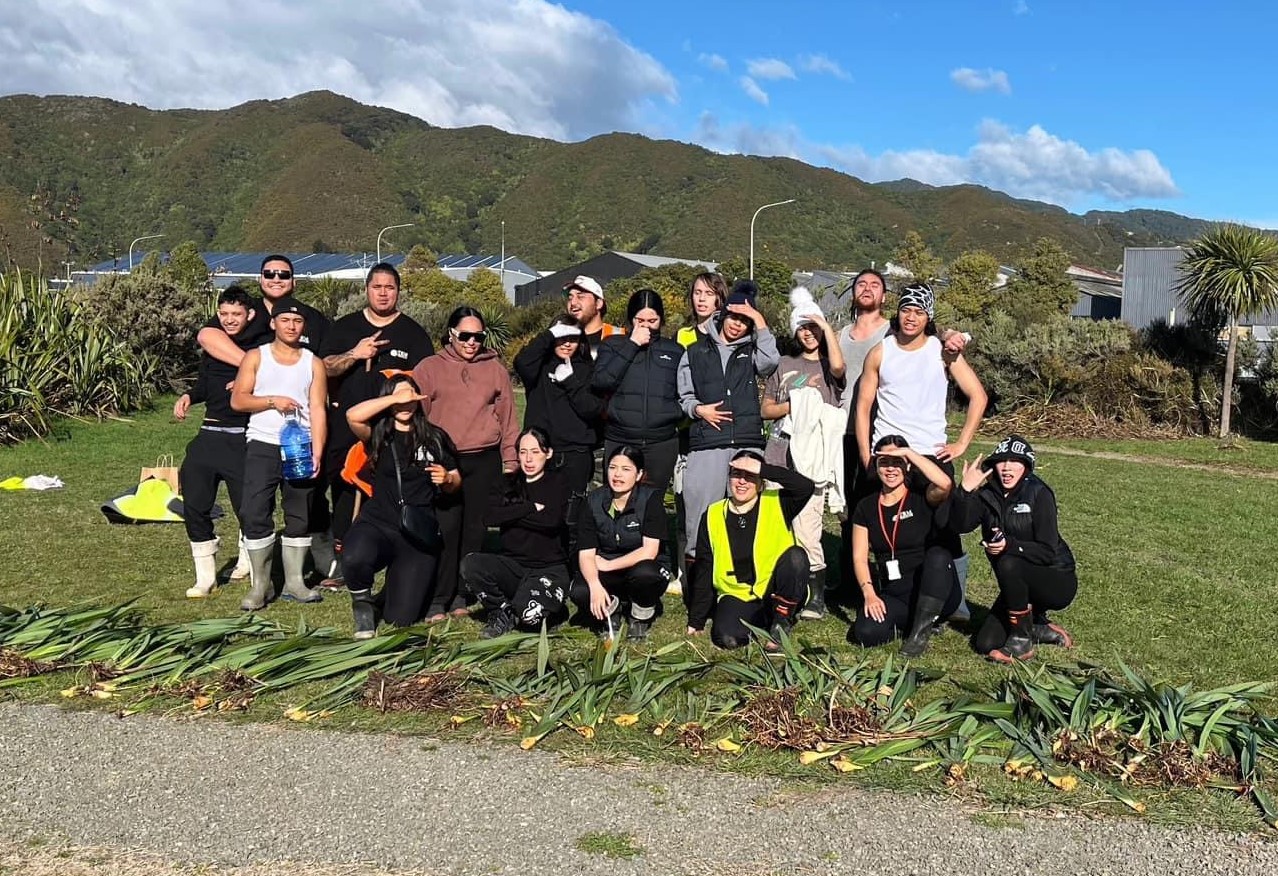 A group of 20 young people outside in front of harakeke flax plants in the sun